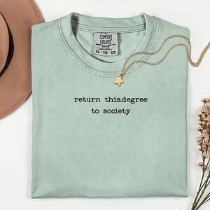May include: A light green t-shirt with the text "return this.degree to society" in black. A gold necklace with a star pendant rests on the shirt. A brown hat is visible in the upper left corner.