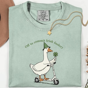 Può includere: T-shirt verde chiaro con la scritta "Off to commit Irish foolery". La maglietta presenta un'oca dei cartoni animati con un cappello verde e un quadrifoglio mentre va in scooter. È visibile anche una collana con pendente a stella dorata.