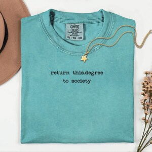 May include: A turquoise Comfort Colors t-shirt with the text "return this.degree to society" in black. A gold star pendant necklace rests on the shirt. A brown hat and flowers are in the background.