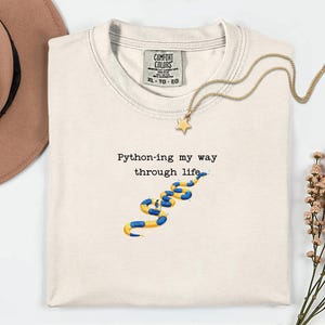May include: Cream-colored t-shirt with the text "Python-ing my way through life" and a blue and yellow snake graphic. A gold necklace with a star pendant and a brown hat are also visible.