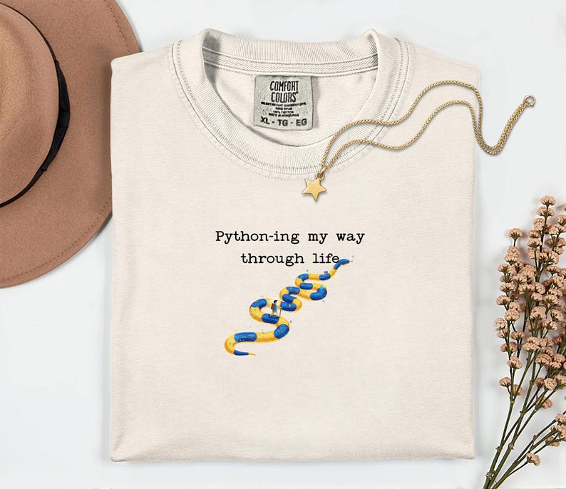 May include: Cream-colored t-shirt with the text "Python-ing my way through life" and a blue and yellow snake graphic. A gold necklace with a star pendant and a brown hat are also visible.