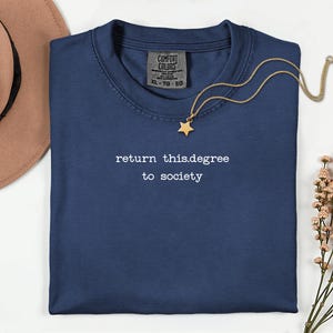 May include: Navy blue crew neck t-shirt with the text "return this.degree to society" in white. A gold necklace with a star pendant rests on the shirt. A brown hat and flowers are in the background.