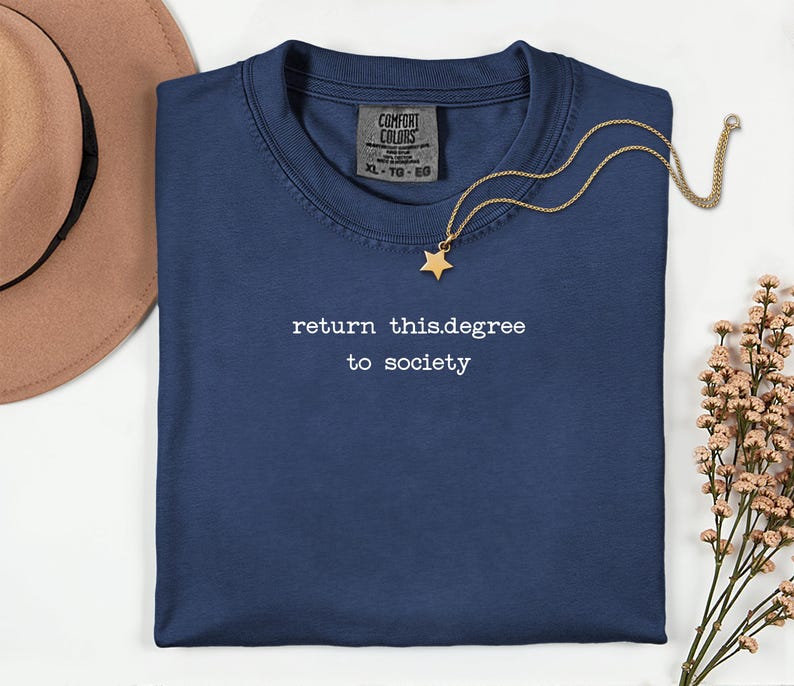 May include: Navy blue crew neck t-shirt with the text "return this.degree to society" in white. A gold necklace with a star pendant rests on the shirt. A brown hat and flowers are in the background.