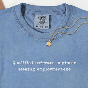 May include: A light blue Comfort Colors t-shirt with the text "Qualified software engineer seeking employment.exe" in white. A gold star pendant necklace rests on the shirt.