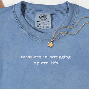 Może przedstawiać: Jasnoniebieska koszulka Comfort Colors z napisem "Bachelor's in debugging my own life". Złoty naszyjnik z gwiazdką spoczywa na koszulce. Koszulka ma okrągły dekolt i metkę XL-TG-EG.