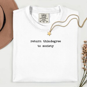 May include: A white t-shirt with the text "return this.degree to society" in black. A gold necklace with a star pendant rests on the shirt. A brown hat and flowers are in the background.