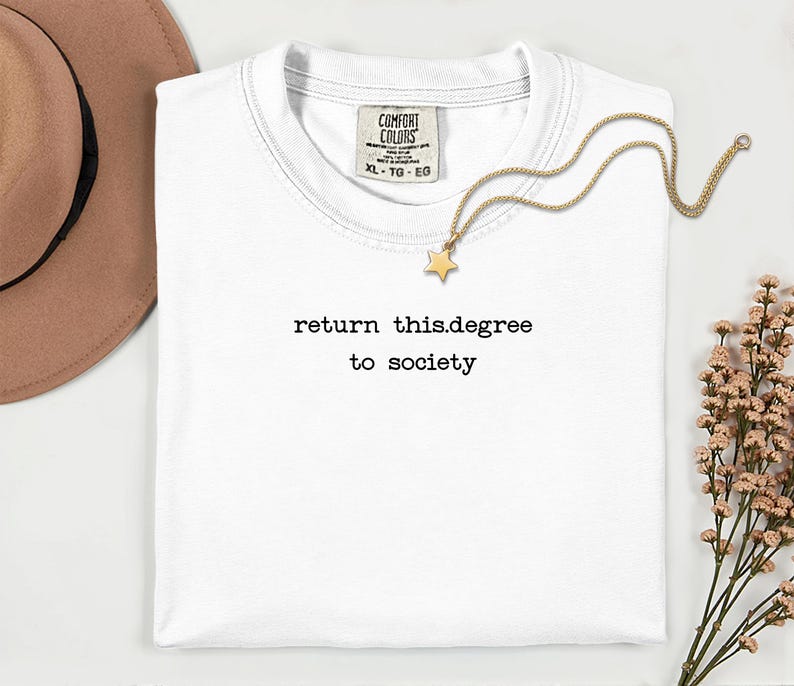 May include: A white t-shirt with the text "return this.degree to society" in black. A gold necklace with a star pendant rests on the shirt. A brown hat and flowers are in the background.