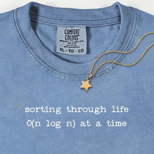 Pode incluir: Camiseta azul clara com o texto branco "sorting through life O(n log n) at a time". Um pingente de estrela dourada pende de uma corrente dourada. A camiseta tem uma etiqueta Comfort Colors.