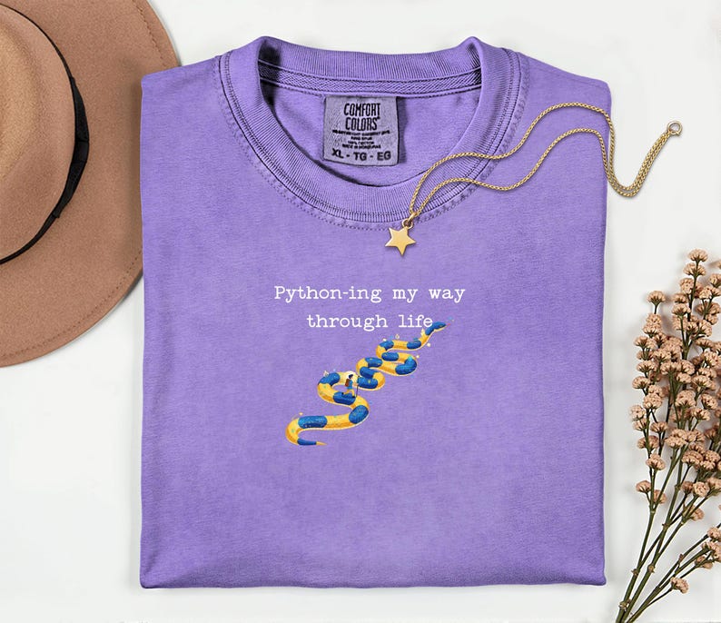 May include: A lilac Comfort Colors t-shirt with the text "Python-ing my way through life" and a snake illustration in yellow and blue. A gold necklace with a star pendant is draped across the shirt.