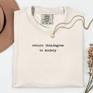 May include: Cream-colored t-shirt with the text "return this.degree to society" in black. A gold necklace with a star pendant rests on the shirt. A brown hat and sprigs of flowers are also in the image.