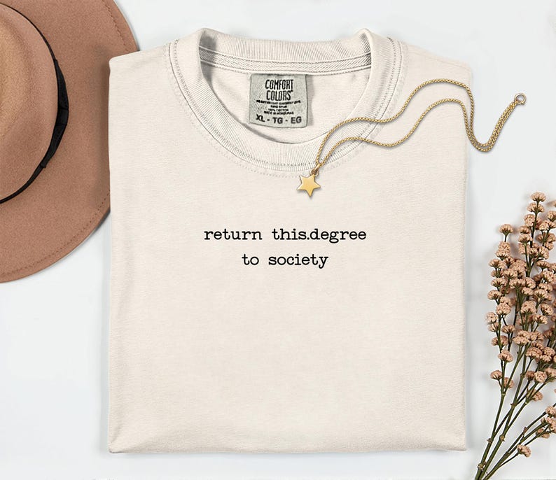 May include: Cream-colored t-shirt with the text "return this.degree to society" in black. A gold necklace with a star pendant rests on the shirt. A brown hat and sprigs of flowers are also in the image.
