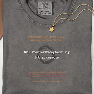 May include: A charcoal grey t-shirt featuring a gold star pendant necklace resting on the collar. The shirt displays white and orange text reading EXCEPTION IN THREAD 'main' java.lang.NullPonexTection, NullPointerException: my job prospects.