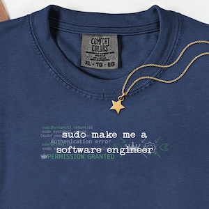 May include: A navy blue t-shirt featuring the white text "sudo make me a software engineer" and "PERMISSION GRANTED" in green, with a gold star pendant necklace resting on the collar. The shirt has a Comfort Colors label showing size XL.