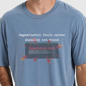 May include: A blue crew neck t-shirt featuring white text that reads "Segmentation fault: career stability not found" above a graphic of a computer error window with the text "Segmentation Fault" and red and yellow digital glitch effects.