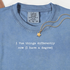 May include: A light blue t-shirt with the text "I Vue things differently now (I have a degree)". A gold star pendant necklace is draped across the shirt. The shirt has a Comfort Colors tag.
