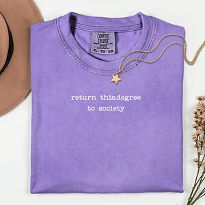 May include: A lilac-colored t-shirt with the text "return this.degree to society" in white. A gold-colored necklace with a star pendant rests on the shirt. A brown hat is visible in the upper left corner.