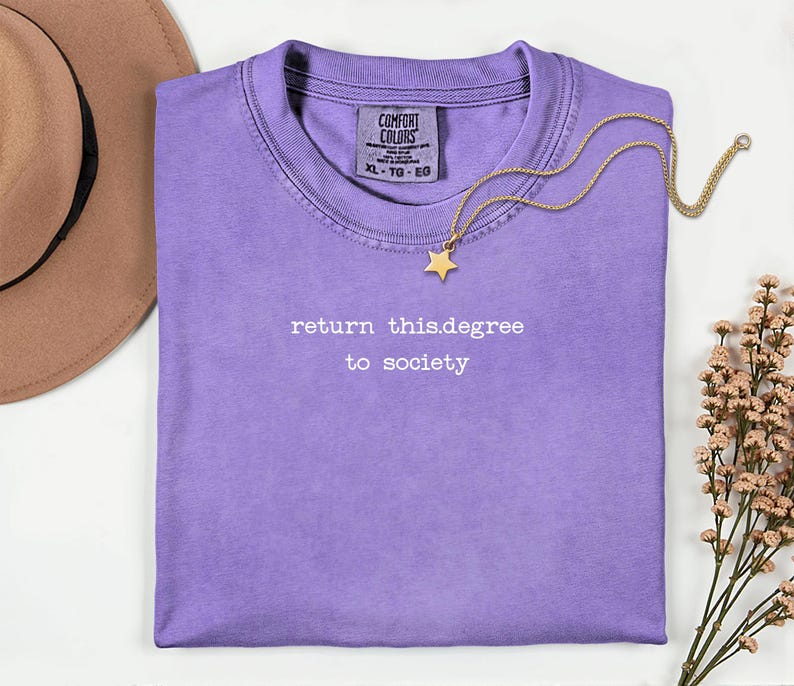May include: A lilac-colored t-shirt with the text "return this.degree to society" in white. A gold-colored necklace with a star pendant rests on the shirt. A brown hat is visible in the upper left corner.