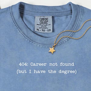 May include: A light blue t-shirt with the text "404: Career not found (but I have the degree)". A gold star pendant necklace rests on the shirt. The shirt has a Comfort Colors tag.