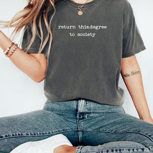 May include: Dark gray t-shirt with the text "return this.degree to society" in white. The shirt is paired with blue jeans and white sneakers. The overall style is casual and relaxed.