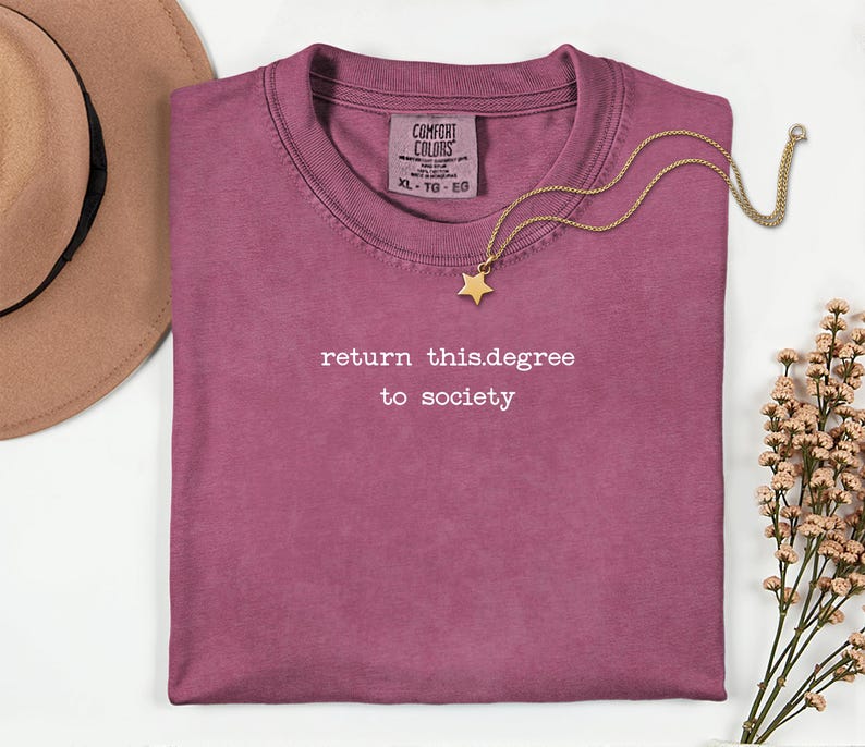May include: A maroon t-shirt with the text "return this.degree to society" in white. A gold necklace with a star pendant rests on the shirt. A tan hat is in the upper left corner, and a small bouquet of flowers is on the right.