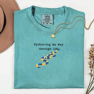 May include: A turquoise Comfort Colors t-shirt with the phrase "Python-ing my way through life" and a snake graphic. A gold necklace with a star pendant is draped across the shirt. A brown hat is visible in the upper left corner.