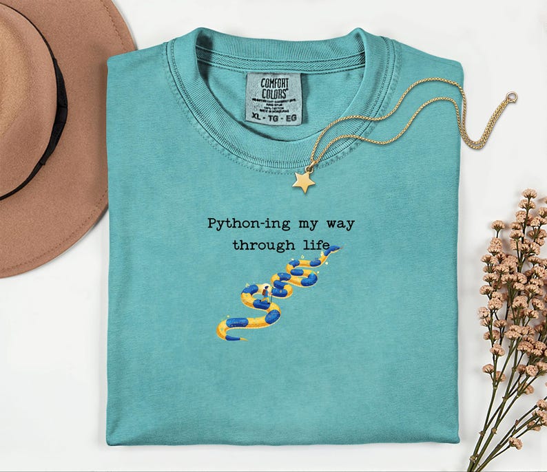 May include: A turquoise Comfort Colors t-shirt with the phrase "Python-ing my way through life" and a snake graphic. A gold necklace with a star pendant is draped across the shirt. A brown hat is visible in the upper left corner.