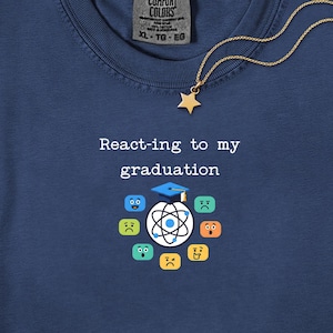 Può includere: Una t-shirt blu navy con il testo bianco React-ing to my graduation sopra un grafico di un atomo con tocco di laurea circondato da varie faccine emoji colorate. Una collana con ciondolo a stella dorata poggia sul tessuto.