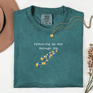 May include: Teal Comfort Colors t-shirt with the text "Python-ing my way through life" and a graphic of a snake made of pool floats. A gold star pendant necklace is draped over the shirt.
