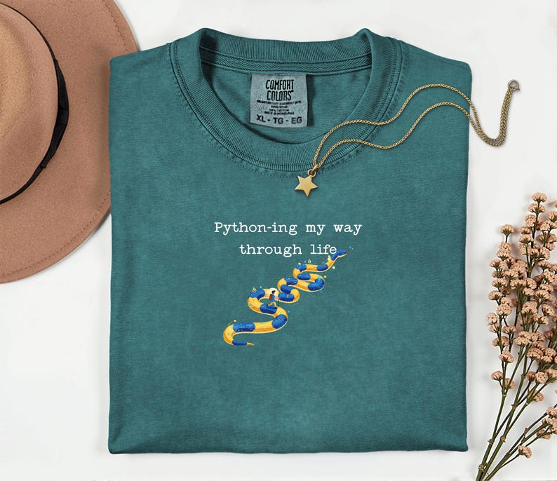May include: Teal Comfort Colors t-shirt with the text "Python-ing my way through life" and a graphic of a snake made of pool floats. A gold star pendant necklace is draped over the shirt.