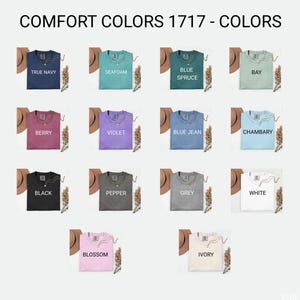 May include: A selection of Comfort Colors 1717 t-shirts in various colors, including True Navy, Seafoam, Blue Spruce, Berry, Violet, Blue Jean, Black, Pepper, Grey, White, Blossom, and Ivory. Each shirt is displayed with a necklace and a hat.