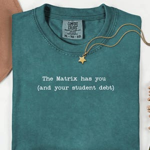 Op de afbeelding: Teal Comfort Colors T-shirt met de tekst "The Matrix has you (and your student debt)". Een gouden sterrenhangerketting is over het shirt gedrapeerd. Het shirt heeft een ronde hals en een label met XL-TG-EG.
