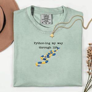 May include: A light green t-shirt with the text "Python-ing my way through life" and a yellow and blue snake illustration. A gold necklace with a star pendant is on the shirt. The shirt has a Comfort Colors tag.