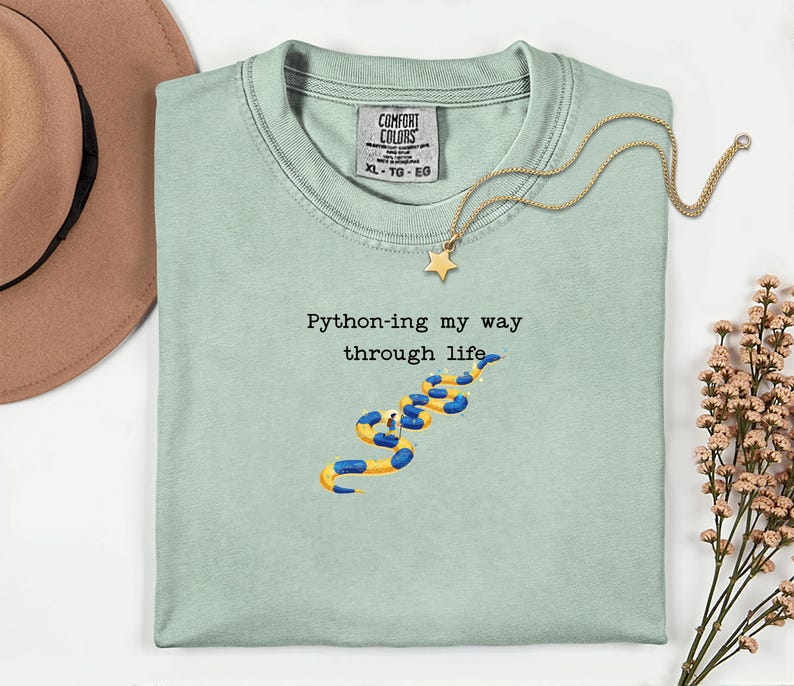 May include: A light green t-shirt with the text "Python-ing my way through life" and a yellow and blue snake illustration. A gold necklace with a star pendant is on the shirt. The shirt has a Comfort Colors tag.