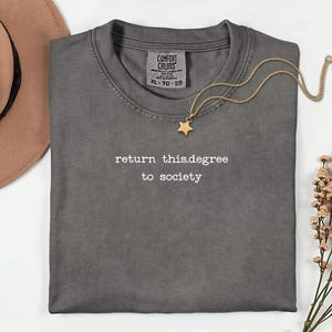 May include: A gray Comfort Colors t-shirt with the text "return this.degree to society" in white. A gold star pendant necklace and a brown hat are also in the image. The shirt is XL.