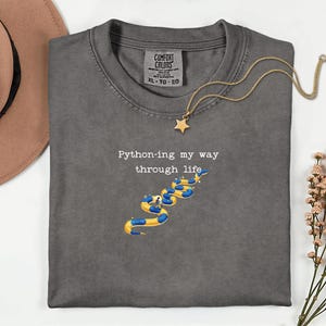 May include: Dark gray t-shirt with the text "Python-ing my way through life" and a snake illustration in yellow and blue. A gold necklace with a star pendant is draped over the shirt. The shirt is a size XL.