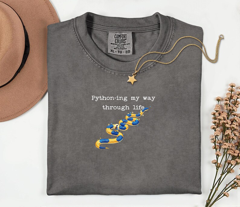May include: Dark gray t-shirt with the text "Python-ing my way through life" and a snake illustration in yellow and blue. A gold necklace with a star pendant is draped over the shirt. The shirt is a size XL.