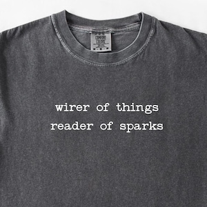 Puede incluir: Camiseta gris oscuro con el texto blanco "wirer of things reader of sparks". La camiseta tiene cuello redondo y la etiqueta de la marca Comfort Colors es visible en el cuello.