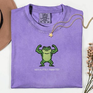 May include: A lilac Comfort Colors t-shirt featuring a cartoon frog flexing its muscles. The frog is green with a white belly and the text "ABSOLUTELY RIBBITED" is below it. A gold star pendant necklace rests on the shirt.
