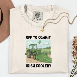Op de afbeelding: Crèmekleurig T-shirt met de tekst "OFF TO COMMIT IRISH FOOLERY" en een afbeelding van een groene tractor die klavers opschept. Een gouden ketting met een sterhanger ligt over het shirt.