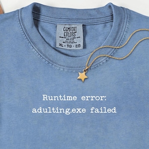 Op de afbeelding: Een lichtblauw Comfort Colors T-shirt met de witte tekst "Runtime error: adulting.exe failed". Een goudkleurige sterhanger hangt aan een fijne ketting. Het shirt heeft een ronde hals en een label met XL-TG-EG.