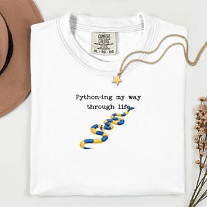 May include: White t-shirt with the text "Python-ing my way through life" and a blue and yellow snake graphic. A gold necklace with a star pendant is draped across the shirt.