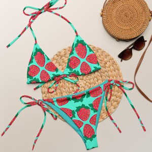 Könnte beinhalten: Ein zweiteiliges Bikini-Set mit Erdbeermuster. Das Bikini-Oberteil und -Unterteil sind hellblau mit einem sich wiederholenden Muster aus roten Erdbeeren. Eine gewebte Strohtasche und Sonnenbrille sind ebenfalls im Bild.