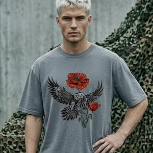 Op de afbeelding: Grijs T-shirt met korte mouwen met een afbeelding van een zwarte adelaar en twee rode klaprozen. Het shirt is gemaakt van een zacht materiaal en heeft een relaxte pasvorm. Het model draagt een groene broek.