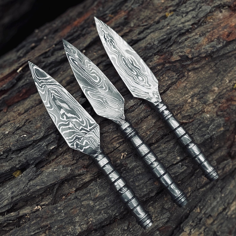 May include: Three handcrafted spearheads with a distinctive patterned blade design. The blades are made of a silver-colored metal, and the handles are a darker, textured metal. The spearheads are displayed on a weathered, dark brown wooden surface.