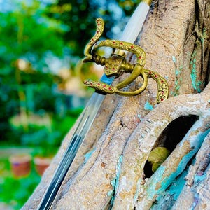 May include: An ornate sword with a polished steel blade and a gold-colored hilt. The hilt features intricate scrollwork and red accents. The white handle contrasts with the gold and steel. The sword is displayed against a tree trunk.