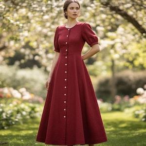 May include: A burgundy-colored dress with a button-down front and short, puffed sleeves. The dress has a full skirt and is worn outdoors in a garden setting. The dress is approximately knee-length.