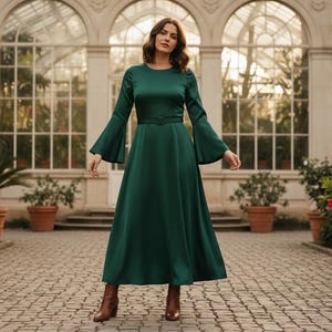 Pode incluir: Um vestido longo verde esmeralda com mangas largas e cintura ajustada. O vestido tem decote redondo e saia fluida. A modelo usa botins castanhos. O fundo inclui um caminho de tijolos, plantas em vasos e uma estufa de vidro.