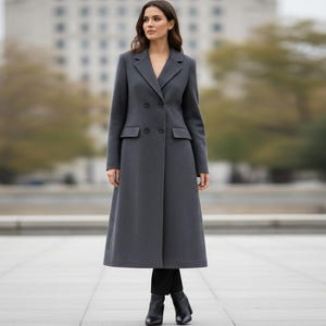 May include: A long, charcoal gray coat with a double-breasted design. The coat features a notched lapel, two front pockets, and a knee-length hem. The person is wearing black boots. The background is a blurred outdoor setting.