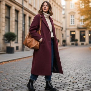 May include: A woman wearing a burgundy long coat, a cream-colored sweater, and blue jeans, carrying a brown leather tote bag. She is wearing black combat boots and walking on a cobblestone street.