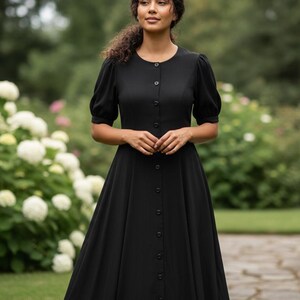 May include: A black dress with a button-down front and short, puffed sleeves. The dress has a fitted bodice and a flowing skirt. The woman is standing in a garden with white and pink flowers.
